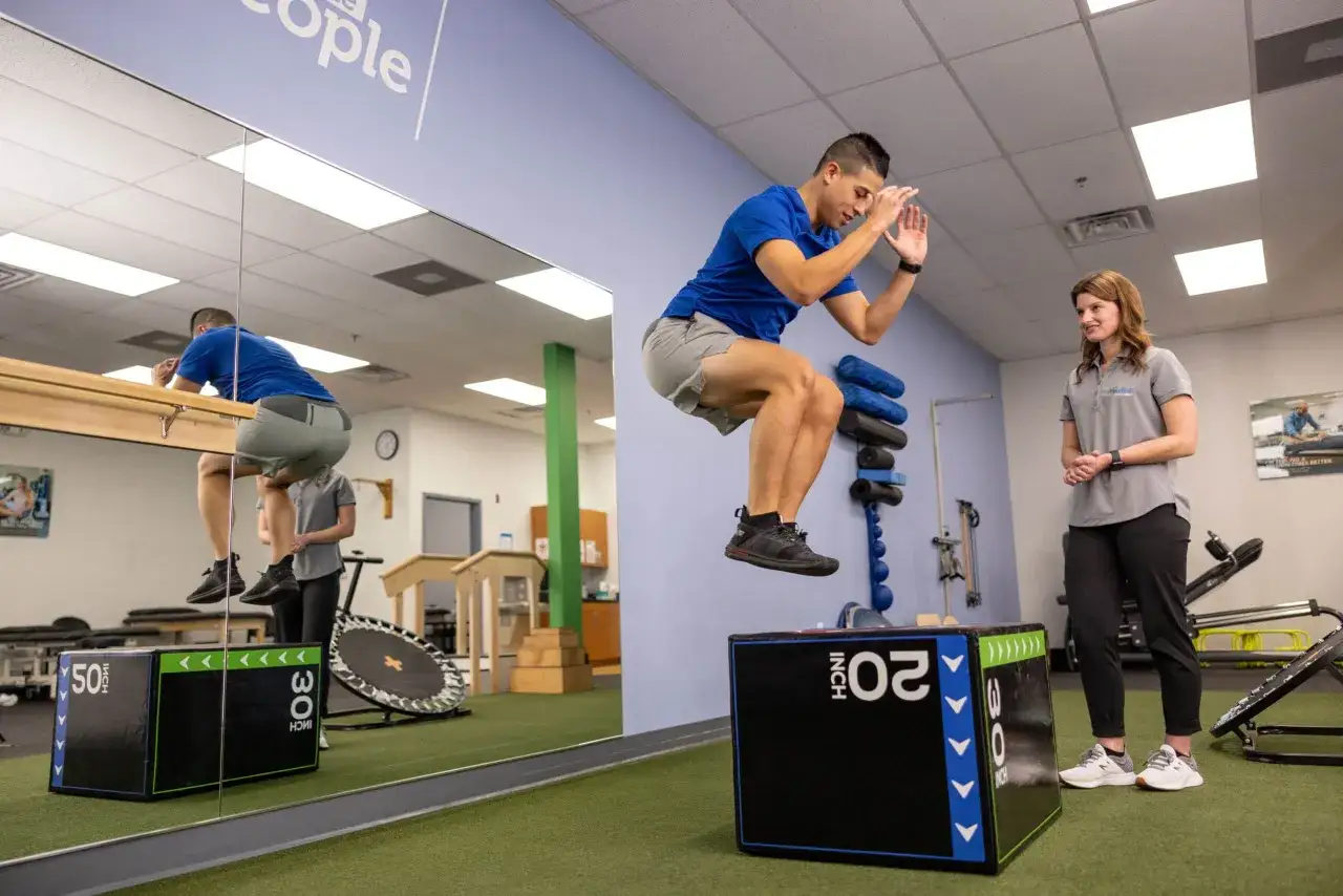 Zawodnik wykonuje wyskok na skrzynię podczas sesji fizjoterapii sportowej. Terapeuta obserwuje jego ruchy.