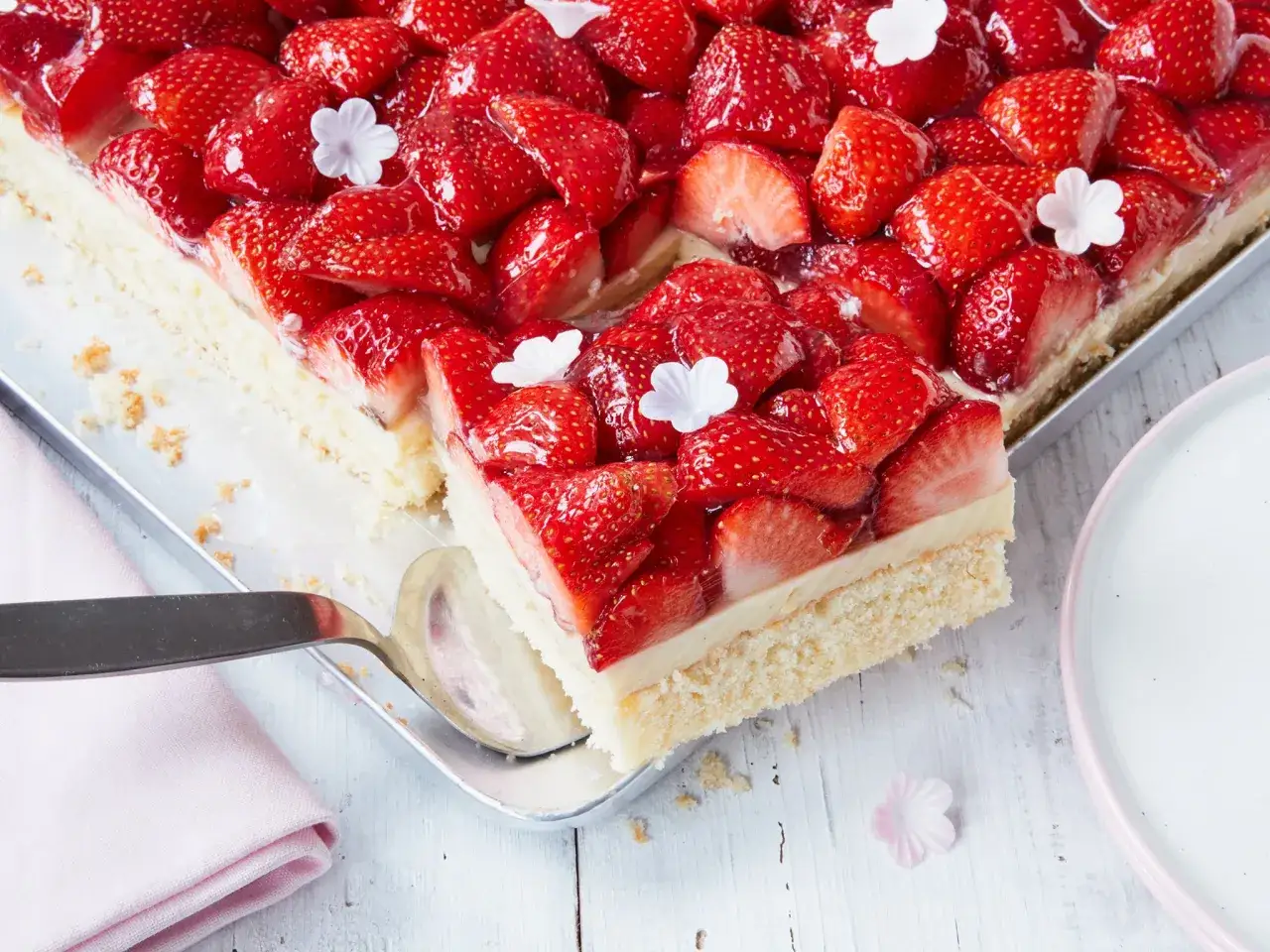 Ein frisch belegter Erdbeerkuchen mit saftigen Erdbeeren und einer cremigen Schicht auf lockerem Boden. Kleine wei&szlig;e Bl&uuml;ten zieren den Kuchen.