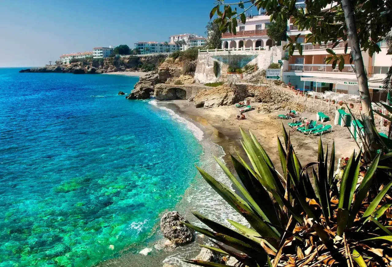 Aguas turquesas y arena dorada en la playa de Nerja, M&aacute;laga. Un para&iacute;so mediterr&aacute;neo con edificios blancos en la costa y vegetaci&oacute;n exuberante.