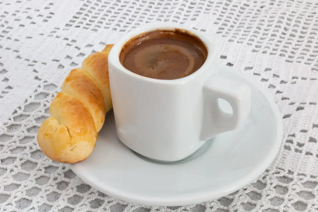 Ein wei&szlig;er Becher mit cremigem mokka griechisch und ein goldbraunes Geb&auml;ck auf einer wei&szlig;en Untertasse, arrangiert auf einer zarten H&auml;keldecke.