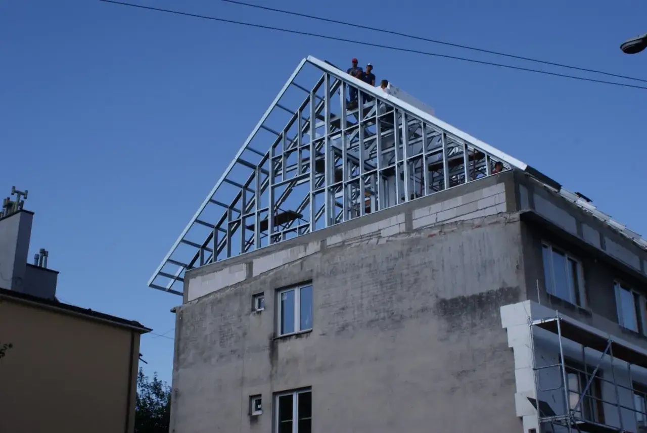 Budowa lekkiej nadbudowy domu. Metalowy szkielet dachu wznosi się nad budynkiem, pracownicy nadzorują postęp prac.