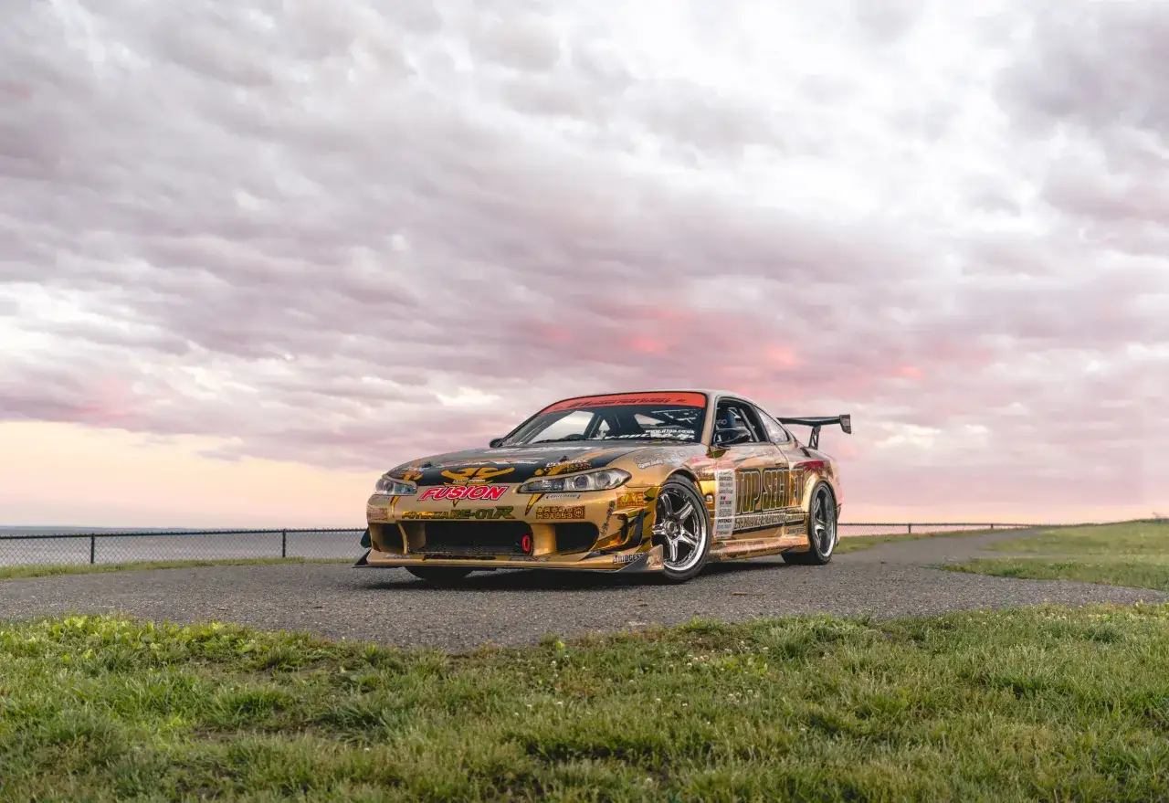 Złoty Nissan S15, jdm japanese drift master, z licznymi naklejkami sponsorów, na tle zachodzącego słońca i chmur.
