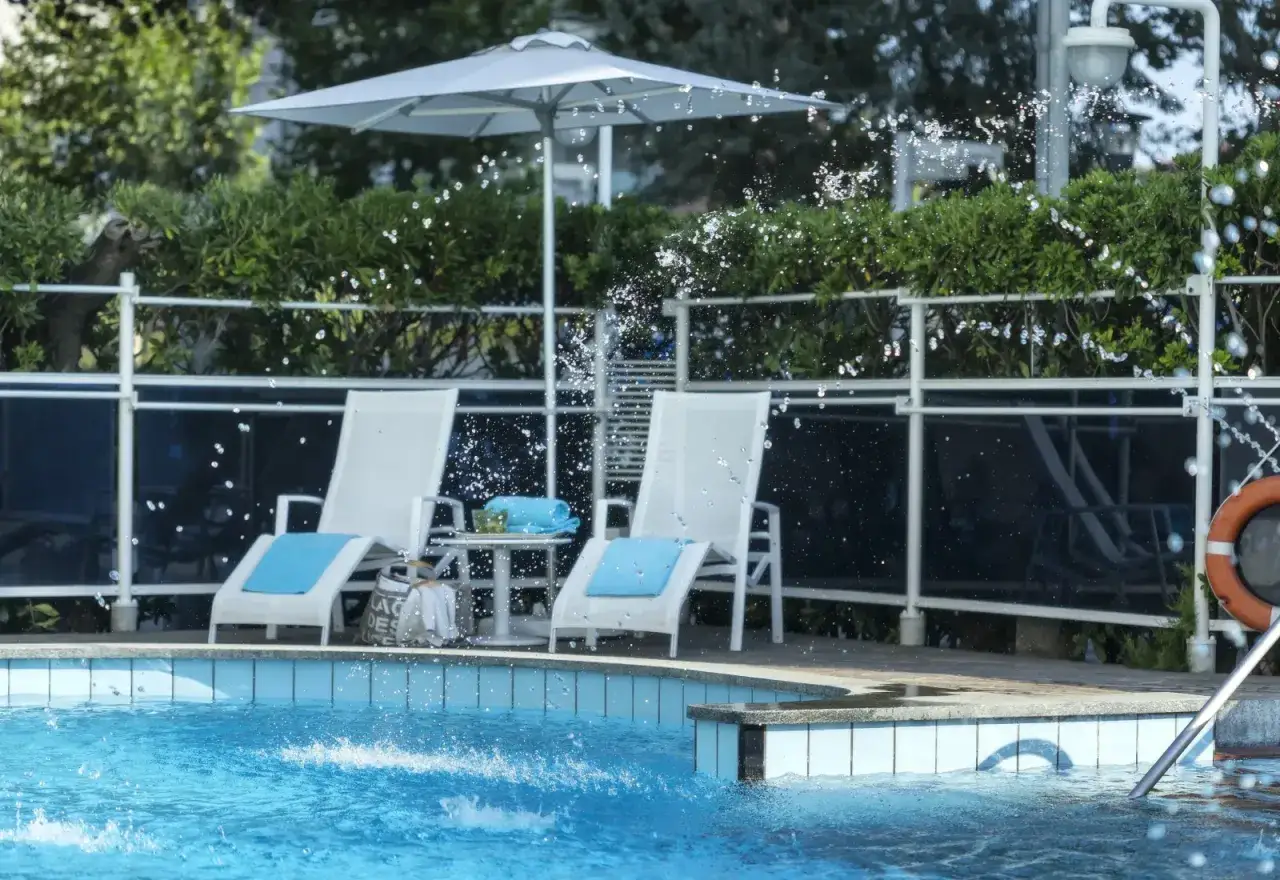 Lettini bianchi con cuscini azzurri e un ombrellone bianco vicino alla piscina del **hotel Abners**. L'acqua schizza vivacemente.