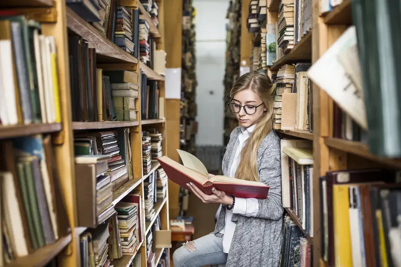 Młoda kobieta w okularach przegląda książkę w bibliotece, otoczona regałami pełnymi **książek, które trzeba przeczytać**.