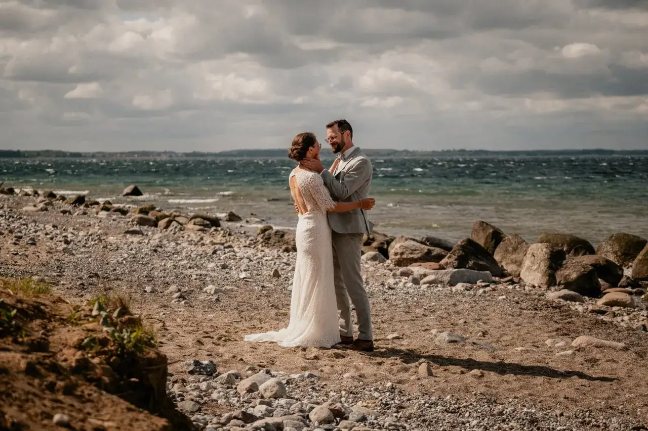 Ein Brautpaar genießt seine Hochzeit am Strand zu zweit. Die Braut trägt ein Spitzenkleid, der Bräutigam einen grauen Anzug. Das Meer und der Himmel sind im Hintergrund zu sehen.