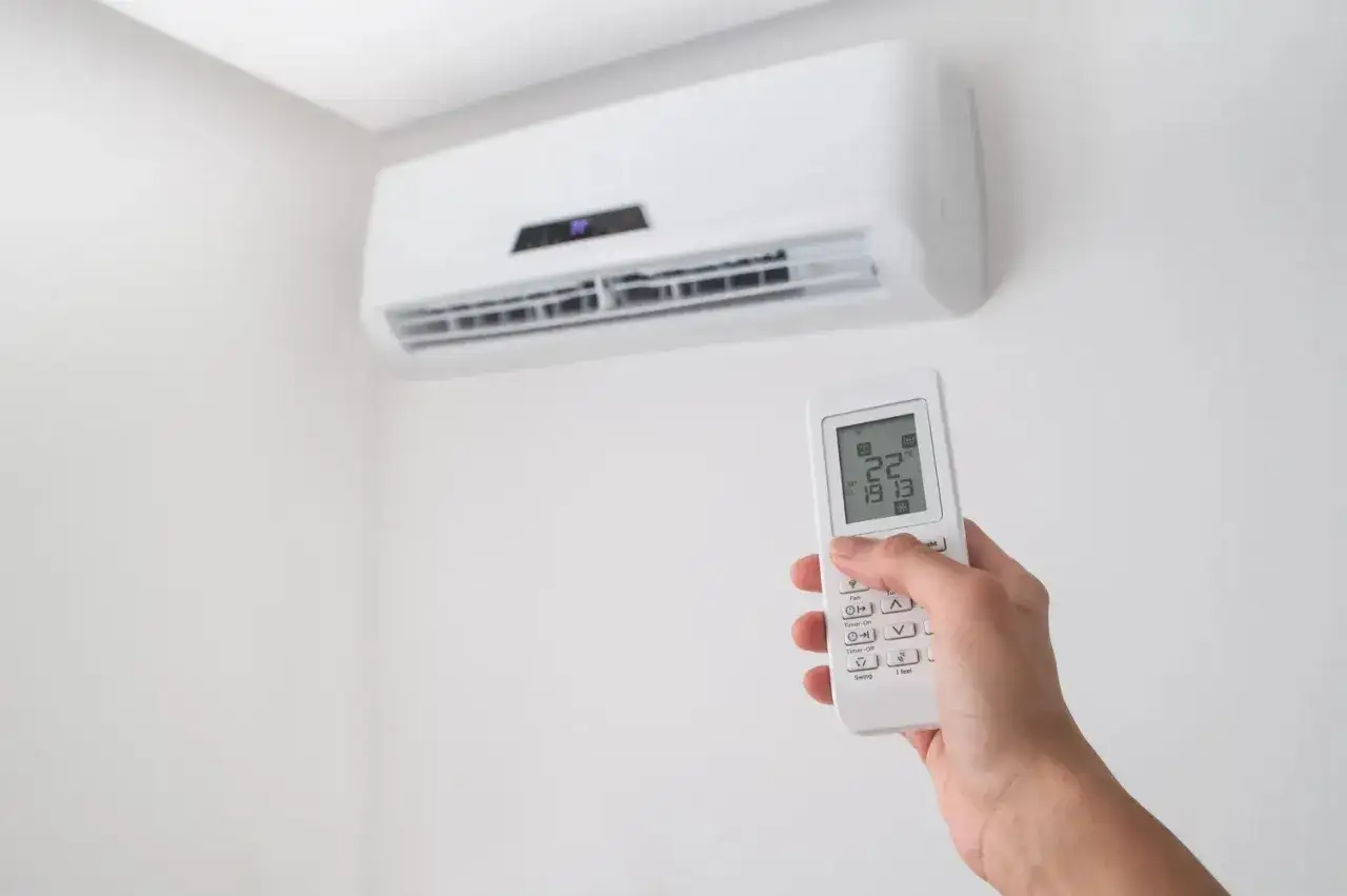 Dłoń trzymająca pilot do klimatyzacji na tle białej ściany z jednostką klimatyzacyjną.