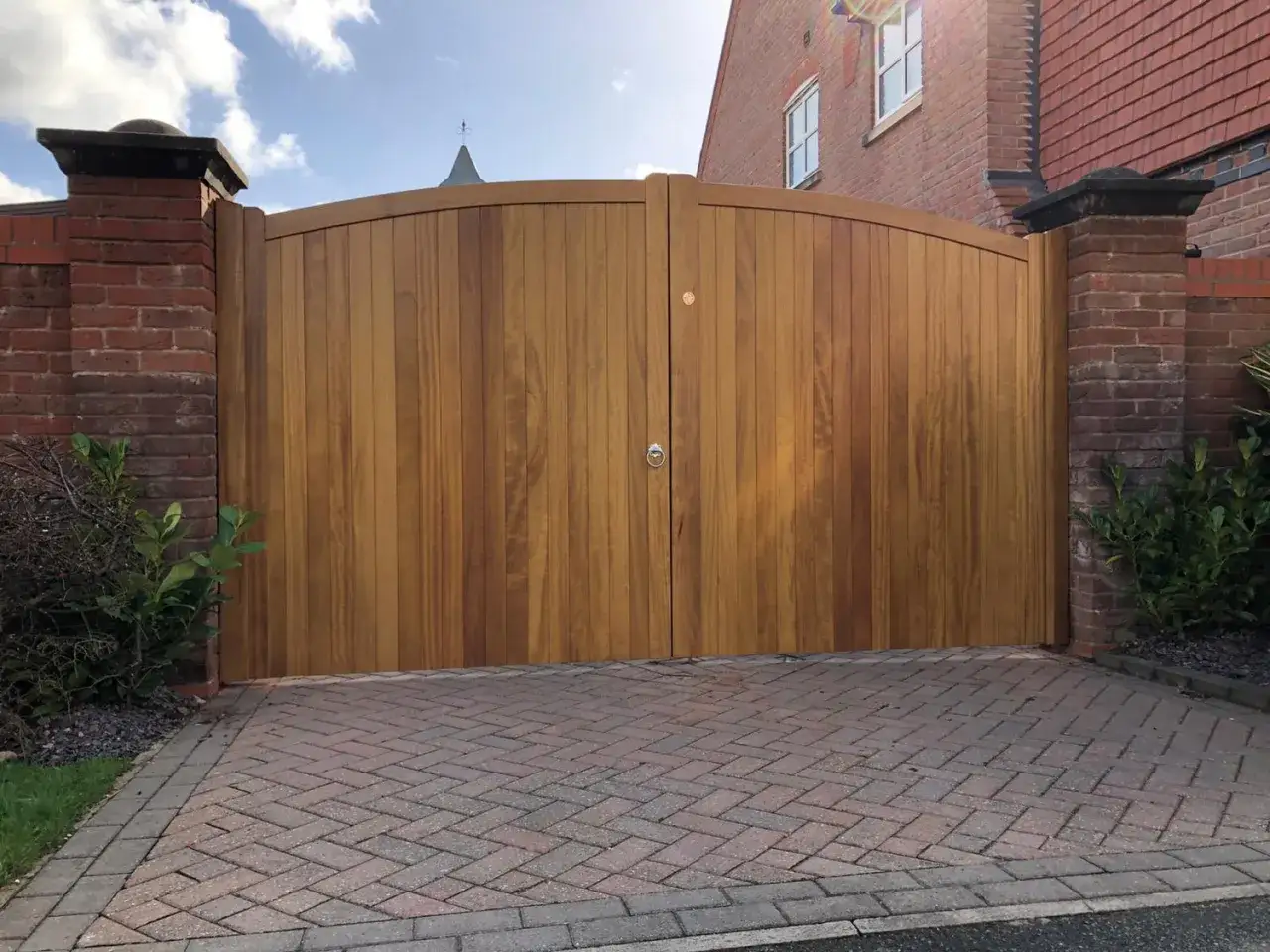 Puerta doble de madera con un cálido color iroko, flanqueada por pilares de ladrillo y un camino de adoquines.