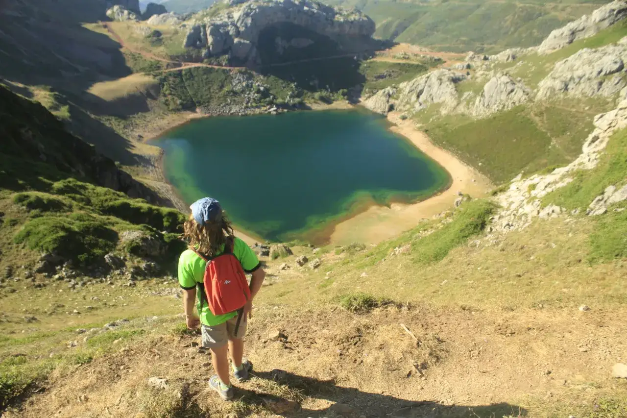 Ni&ntilde;o con mochila naranja explora las **rutas circulares en Somiedo**, admirando un lago turquesa desde la ladera.