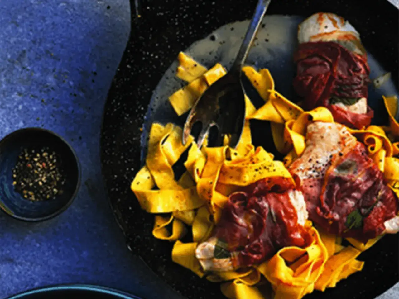 Tacchino zawinięty w prosciutto, podany z szerokim makaronem tagliatelle i posypany pieprzem.