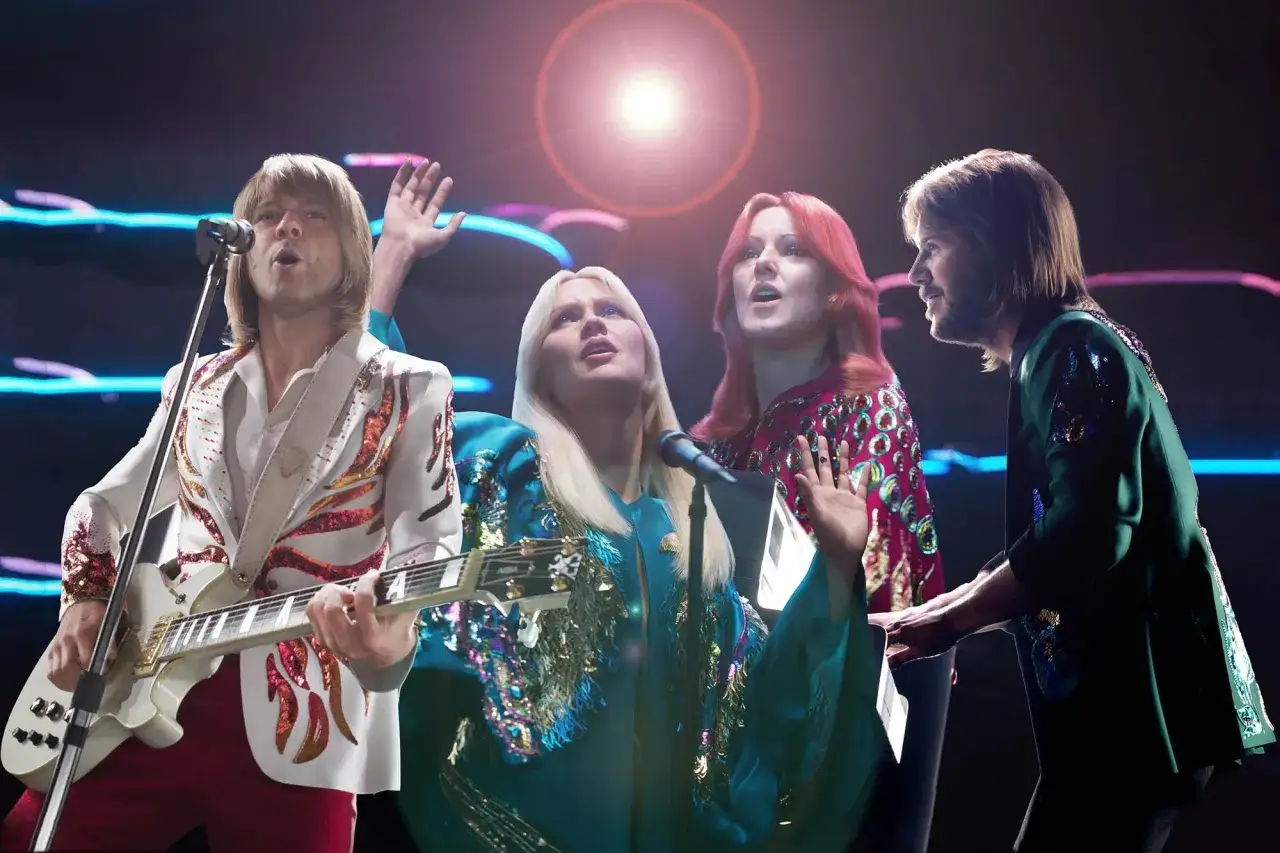 Zesp&oacute;ł ABBA na scenie w błyszczących strojach. Agnieszka śpiewa, Bj&ouml;rn gra na gitarze, a Anni-Frid i Benny towarzyszą im wokalnie.