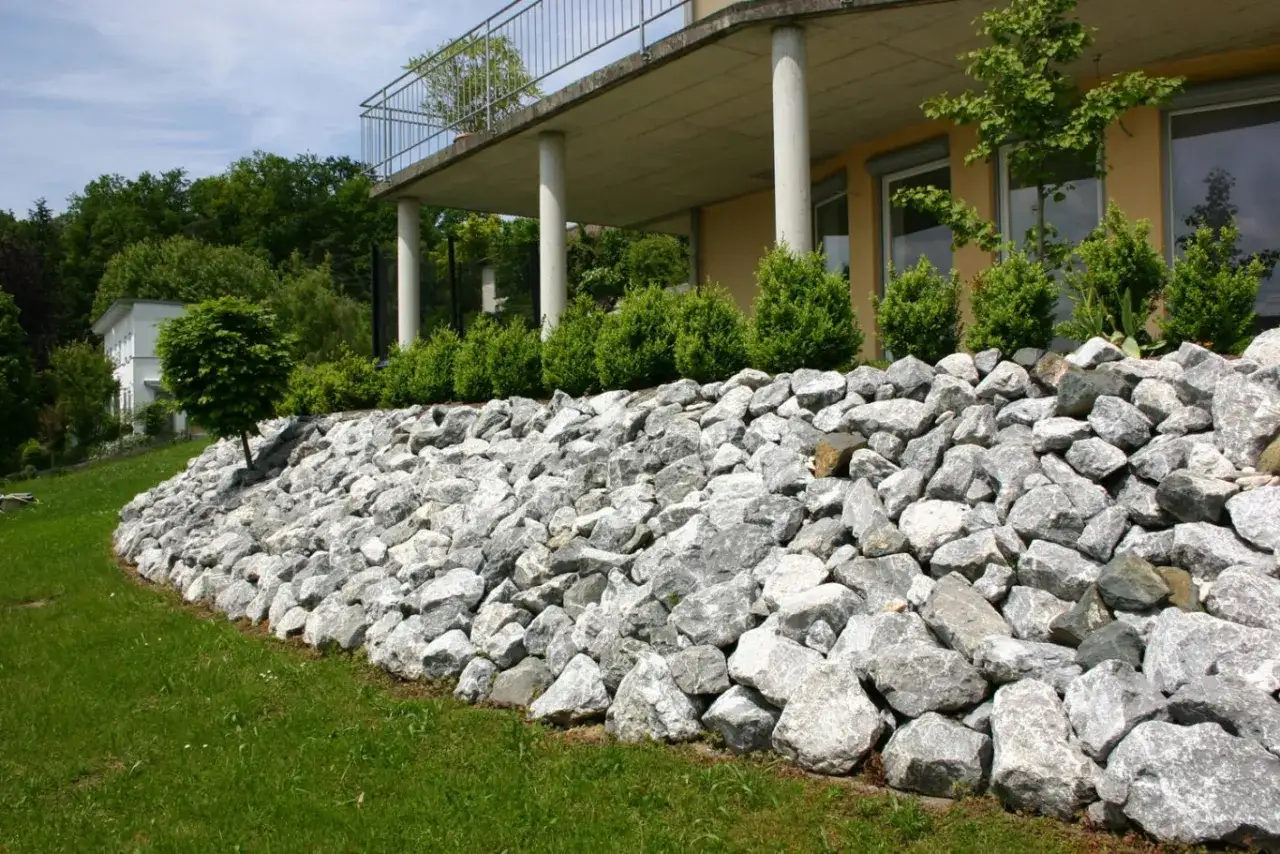 Gro&szlig;e Steine sichern den Hang. Dahinter eine Terrasse mit S&auml;ulen und B&uuml;schen.
