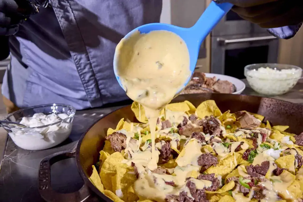 Un cuchar&oacute;n azul vierte un cremoso queso que se derrite sobre nachos con carne.