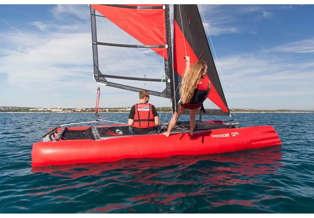 Un couple profite d'une journée ensoleillée sur un catamaran rouge vif. La femme est debout, s'accrochant au mât, prête à utiliser le trapèze.