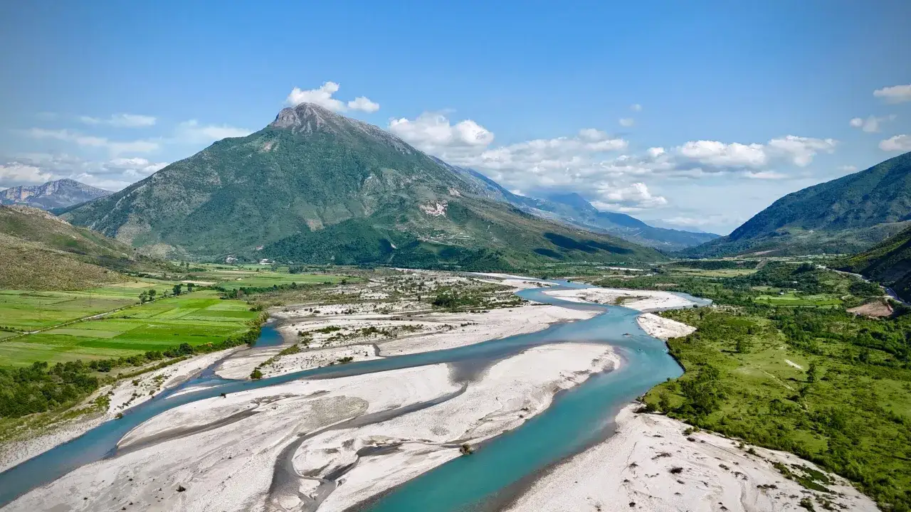 Ein malerischer Fluss schl&auml;ngelt sich durch eine gr&uuml;ne Landschaft in Albanien, mit einem majest&auml;tischen Berg im Hintergrund.