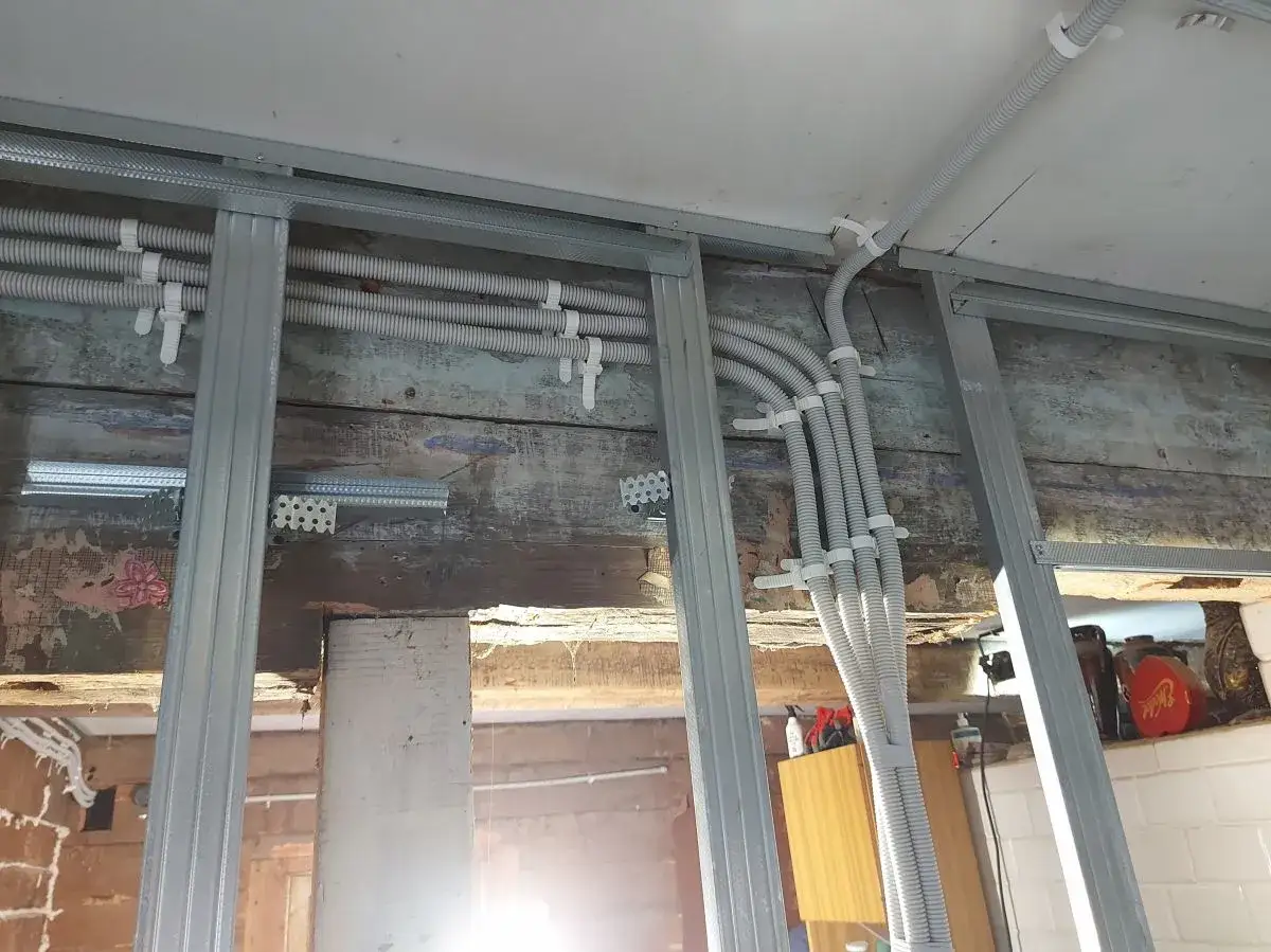 Metalowa konstrukcja ścianki działowej z ułożonymi w peszlach przewodami elektrycznymi. Widać stare, drewniane deski i cegły.