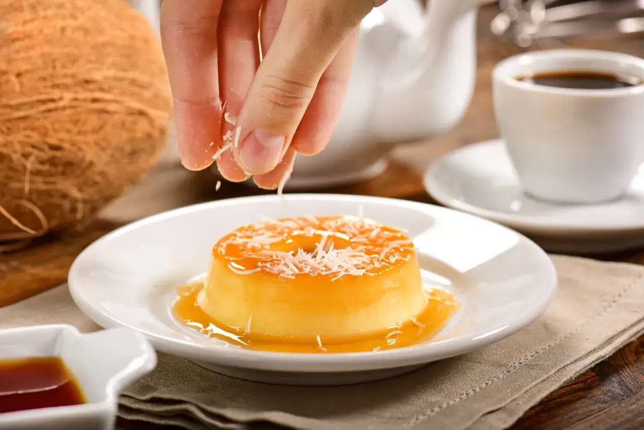 Un flan au lait de coco sans four, garni de noix de coco râpée, est servi sur une assiette blanche avec un coulis de caramel. Une tasse de café est en arrière-plan.