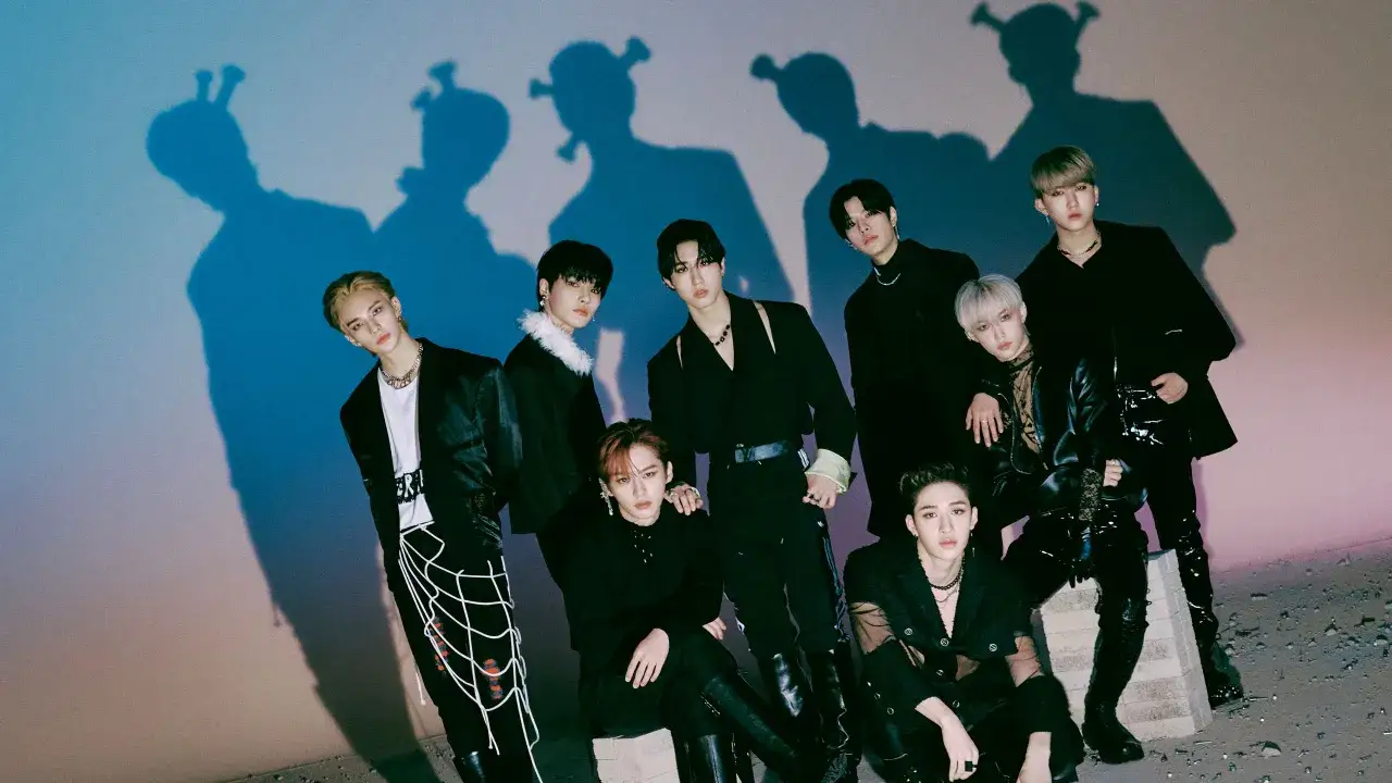 Die Mitglieder von Stray Kids posieren in stylischer schwarzer Kleidung vor einem blauen und rosa Hintergrund, ihre Schatten werfen interessante Formen.