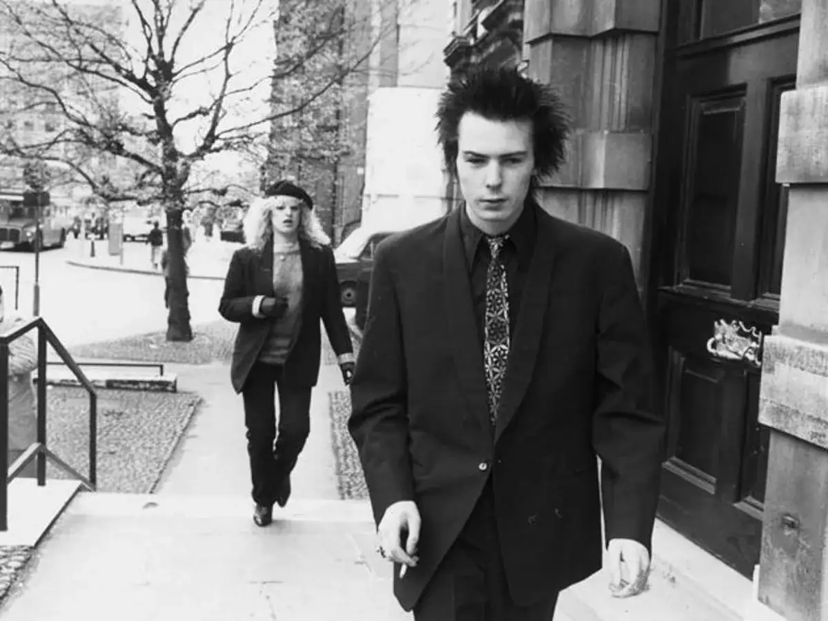 Sid Vicious und Nancy Spungen gehen eine Treppe hinunter. Sid, mit zerzaustem Haar und Anzug, scheint in Gedanken versunken, vielleicht erf&uuml;llt er seine sid wishes.
