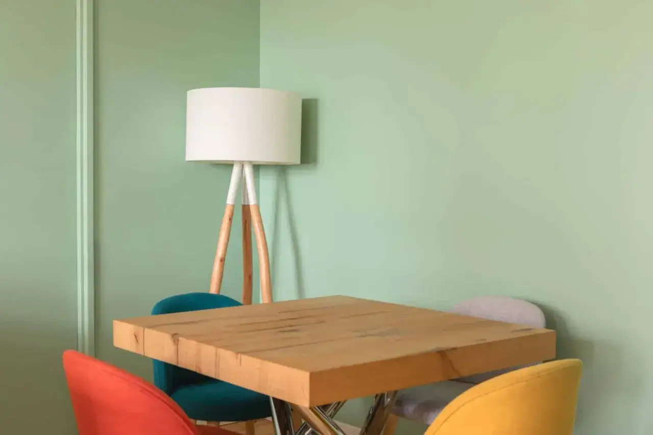 Un coin repas avec une table en bois, des chaises colorées et un lampadaire, le tout sur un mur vert pastel. Quel type de peinture pour les murs ? Une peinture mate pour un rendu doux.