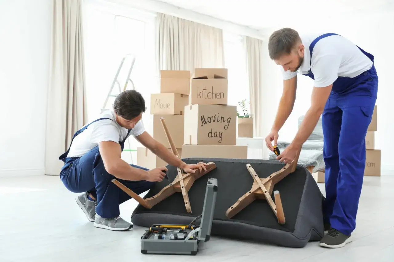 Deux déménageurs s'occupent de comment démonter un canapé fixe. Des cartons de déménagement sont empilés derrière eux.