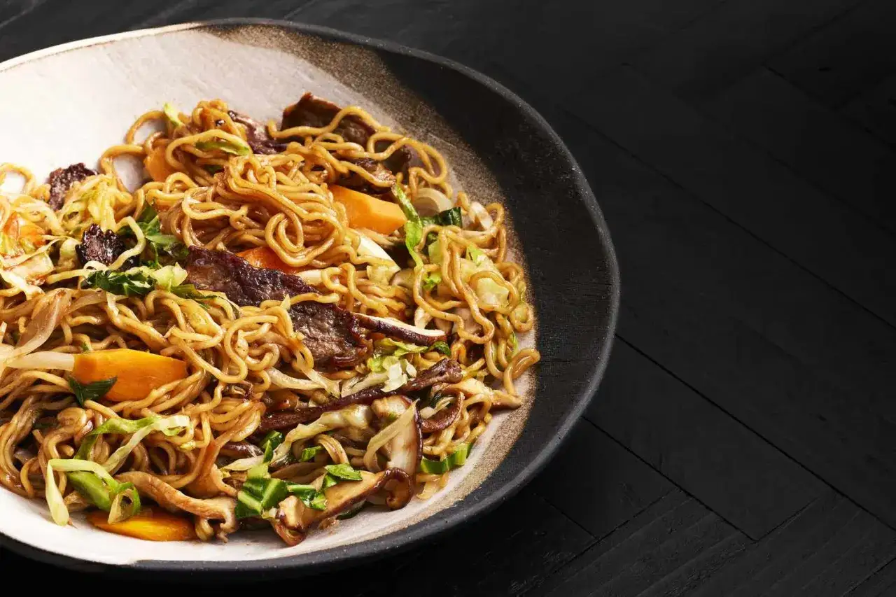 Eine Schüssel mit Yaki Soba, Nudeln mit Rindfleisch, Kohl, Karotten und Pilzen, serviert auf einem dunklen Holztisch.