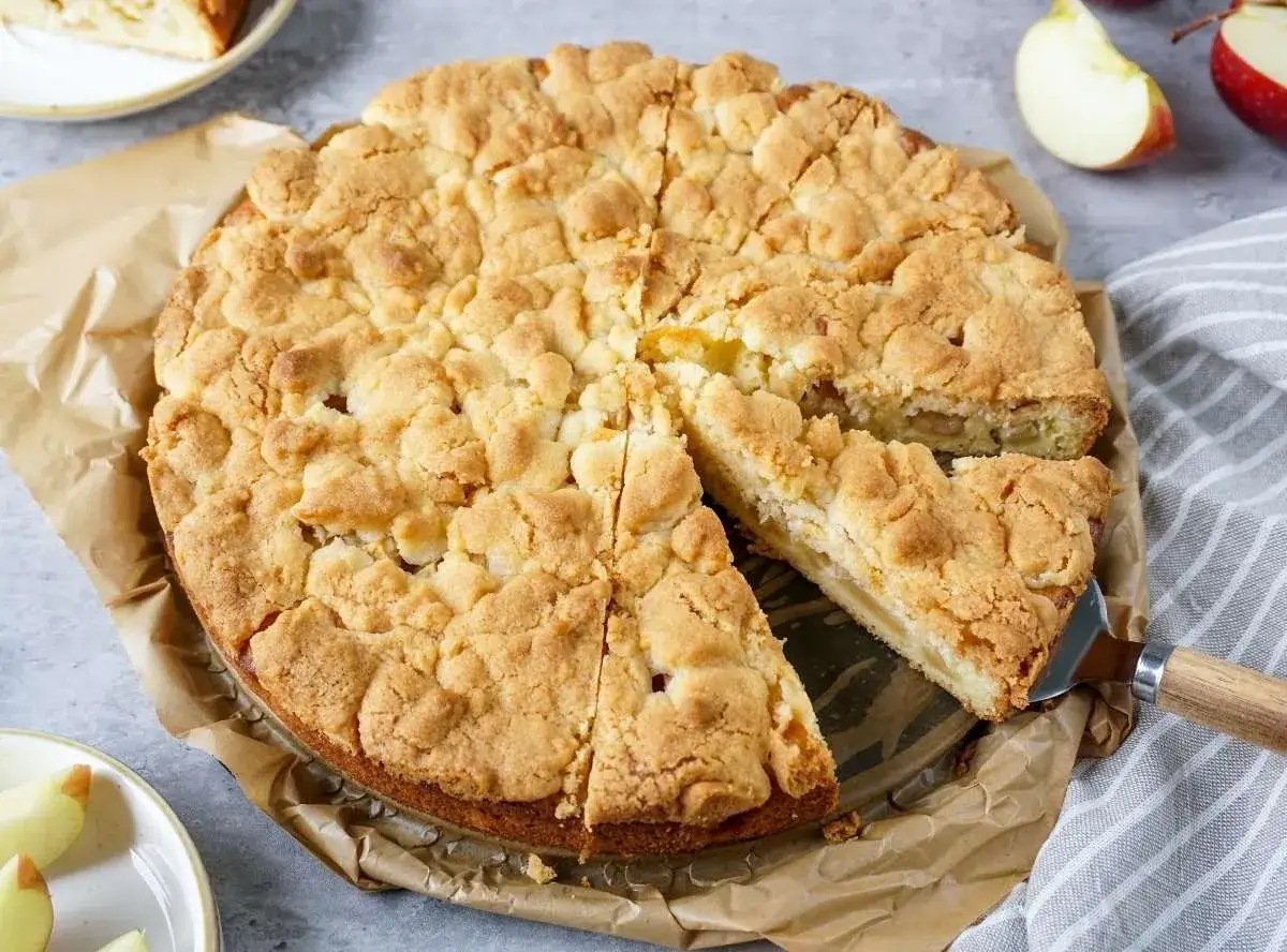 Ein goldbrauner Apfelkuchen mit Streuseln, ein Stück wird gerade herausgenommen.
