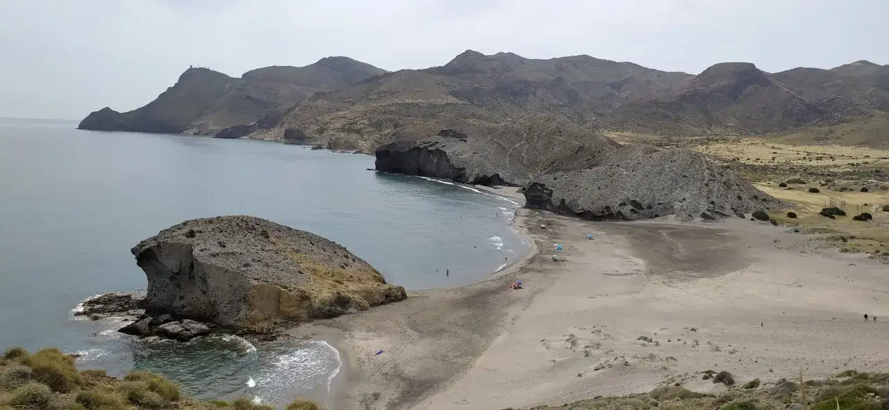 Costa rocosa con el faro de los genoveses en la distancia. Una playa de arena gris con algunas personas y sombrillas se curva hacia el mar azul.