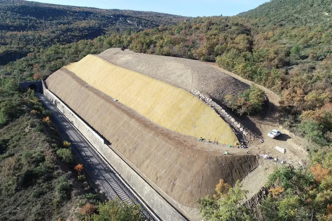 Terreno en ladera con malla y hormigón proyectado, junto a vías de tren y túnel.