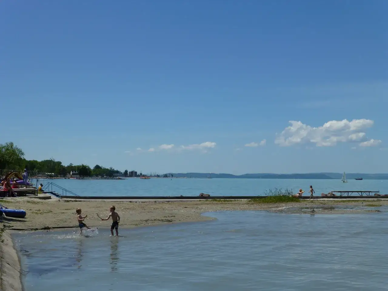 Dzieci bawią się w płytkiej wodzie na plaży. Balaton zachwyca czystością wody, w oddali widać żaglówki.