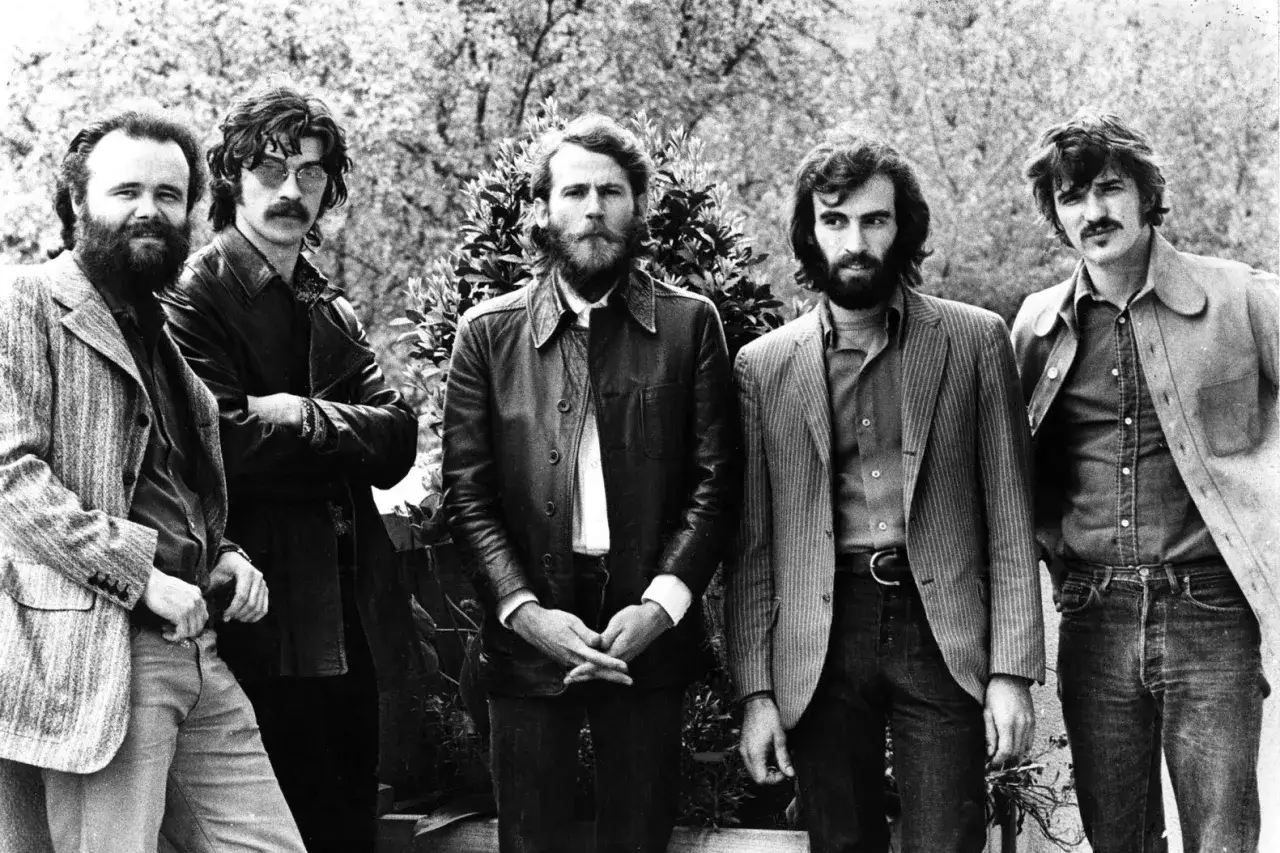 Five men stand in a line, posing for a photo. The man on the far left has a beard and wears a patterned jacket. The man next to him sports a mustache and sunglasses. The other three men have beards and wear various jackets and shirts. They look like th...