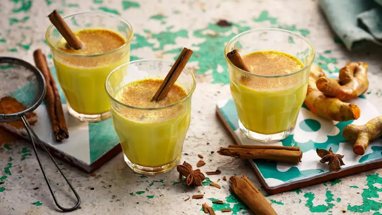 Trzy szklanki rozgrzewającego, złotego mleka z cynamonem i kurkumą, obok świeżych korzeni kurkumy i gwiazdek anyżu.