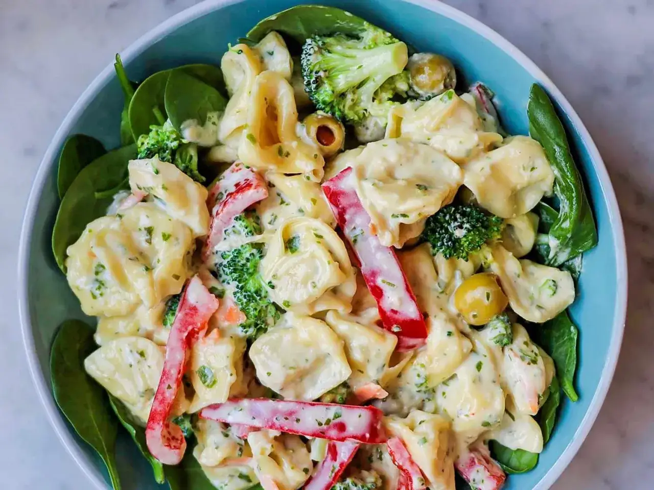 Cremiger Tortellini Salat mit Brokkoli, Paprika und Oliven in einer blauen Sch&uuml;ssel, garniert mit frischem Spinat.