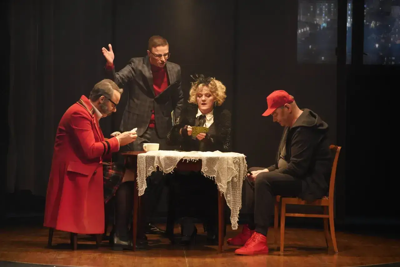 Scena z dramatu Mrożka: cztery postacie przy stole, grają w karty. Jedna z nich, w czerwonym płaszczu, czyta list.