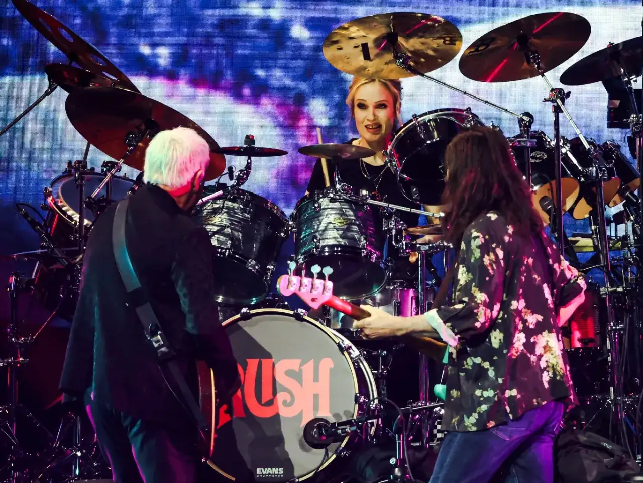 Koncertowy **Rush w Polsce** z perkusistką za bębnami i muzykami grającymi na gitarze basowej i elektrycznej.