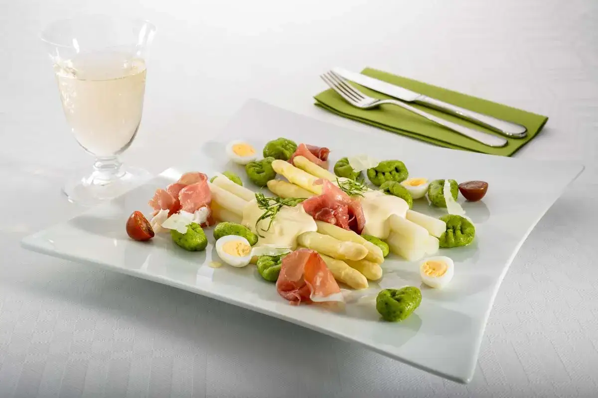 Spargel mit Hollandaise, garniert mit Schinken, Eiern und gr&uuml;nen Kl&ouml;&szlig;chen. Ein Glas Wei&szlig;wein steht daneben.