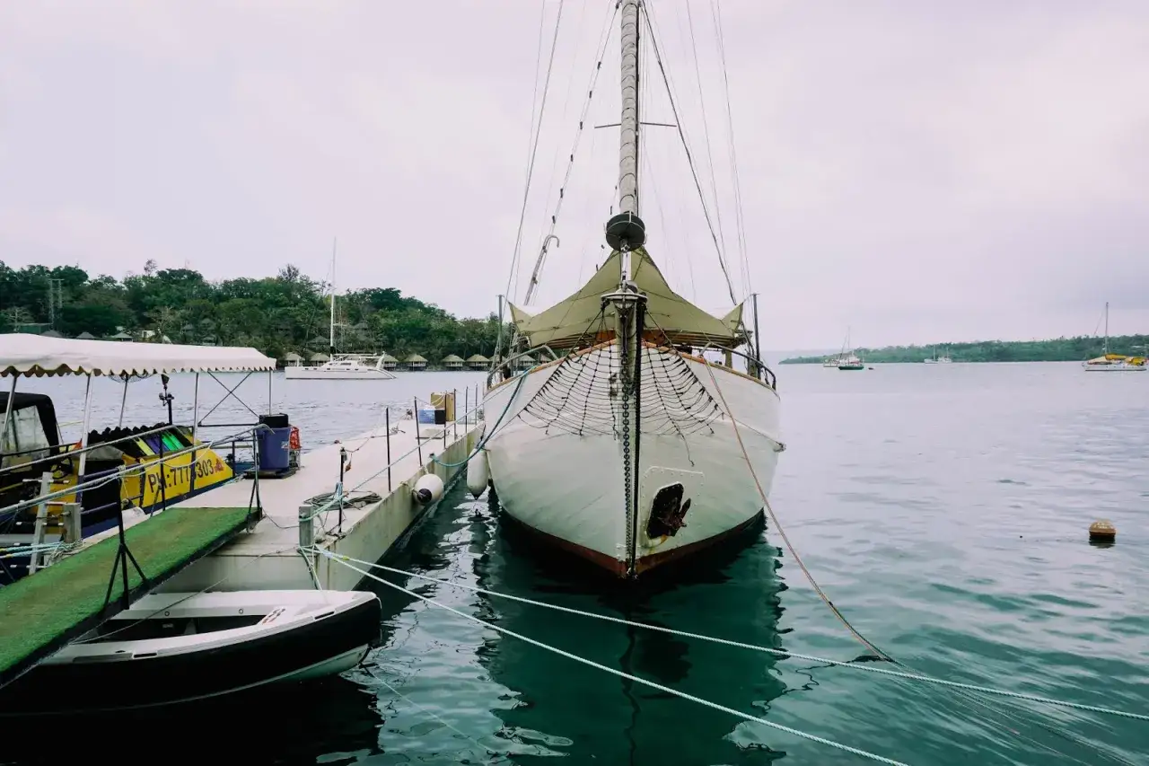 Biały żaglowiec cumuje przy pomoście na wyspach Vanuatu. W tle widać inne łodzie i tropikalną zieleń.