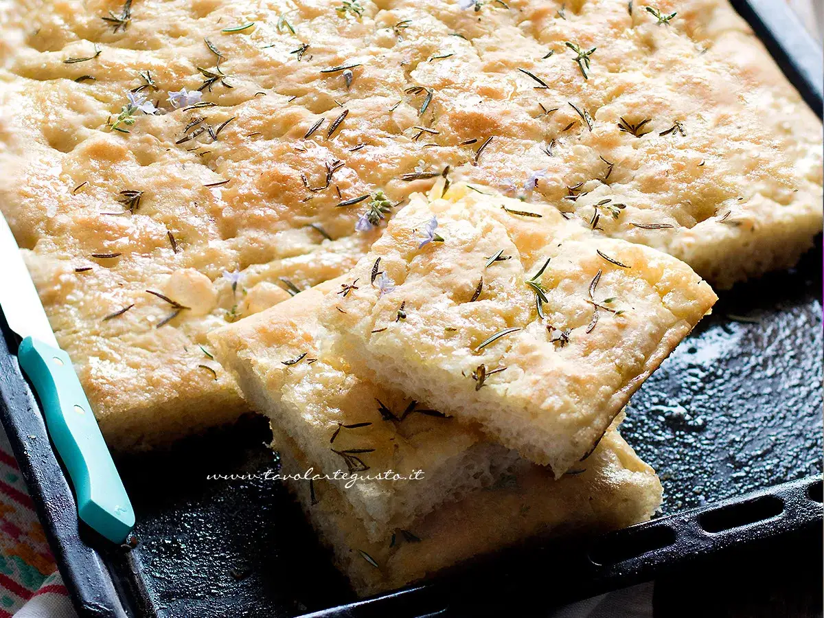Focaccia senza glutine dorata e fragrante, guarnita con rosmarino e piccoli fiori viola, pronta per essere gustata.
