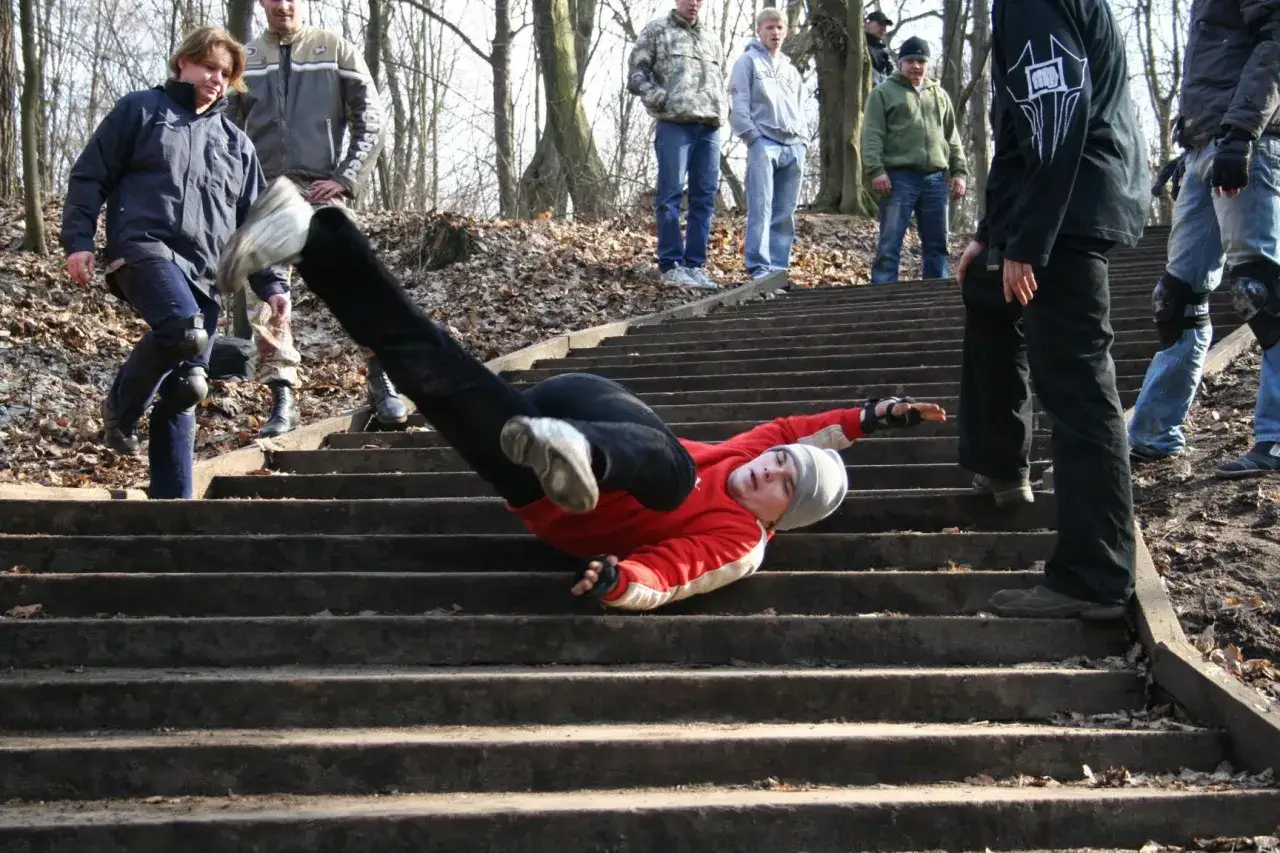 Chłopak w czerwonej bluzie wykonuje kaskaderski upadek po schodach, niczym the ultimate stuntman. Widzowie obserwują jego wyczyn.