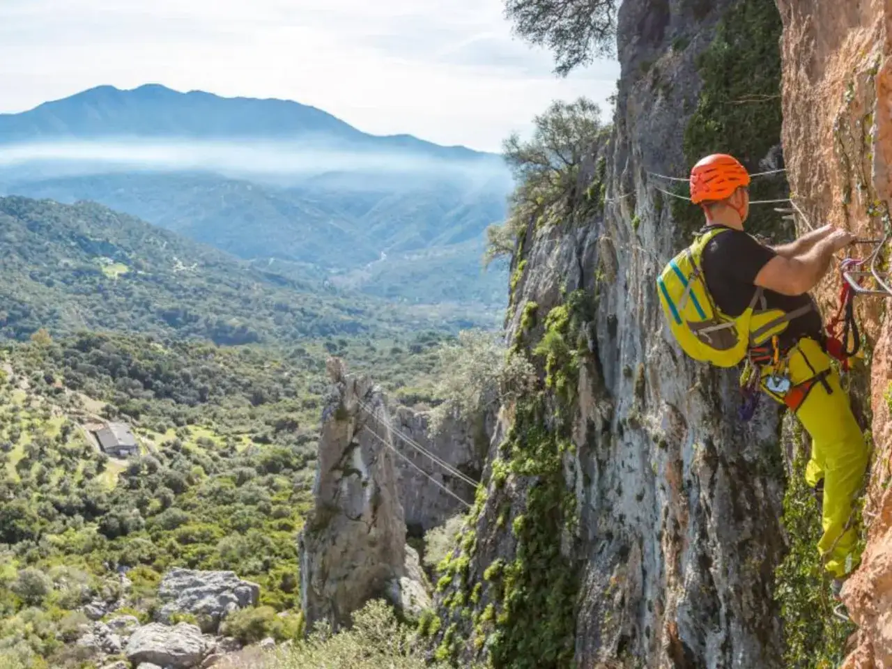 Hombre en equipo de seguridad asciende por una pared rocosa, disfrutando de un paisaje montañoso. Esto es una vía ferrata.