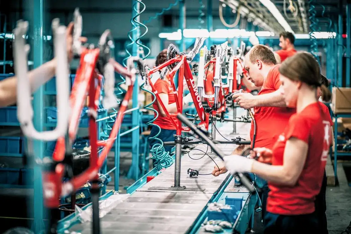 Pracownicy w fabryce Romet składają rowery. Czerwone ramy rowerowe wiszą na linii produkcyjnej, a pracownicy montują części.