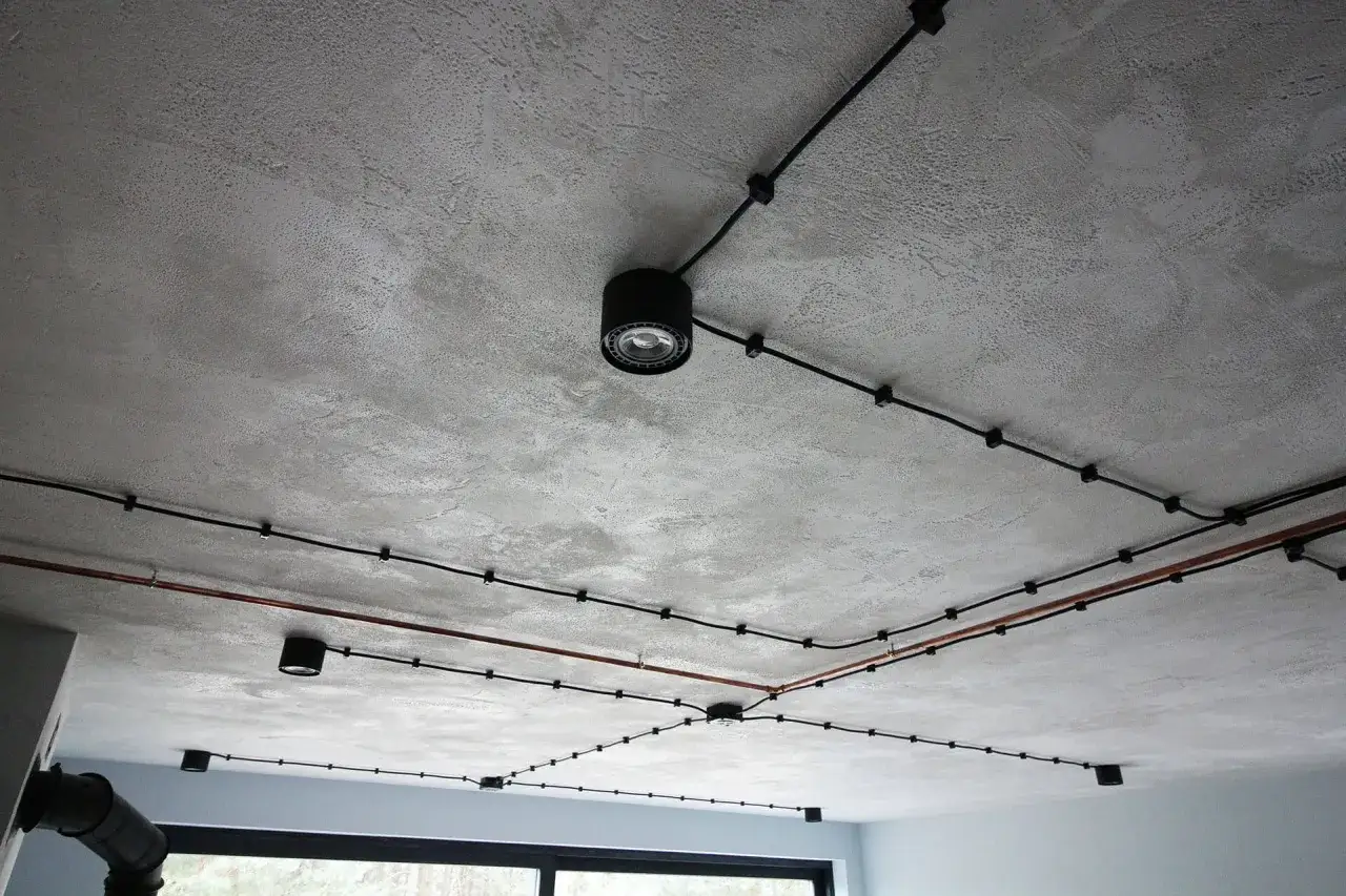 Surowy, betonowy sufit w stylu loft z widocznymi czarnymi przewodami i oprawami oświetleniowymi.