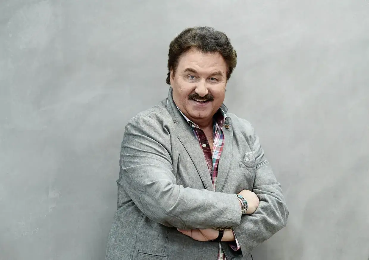 Uśmiechnięty Krzysztof Krawczyk, którego wiek zdradza doświadczenie, w szarym garniturze i kolorowej koszuli.