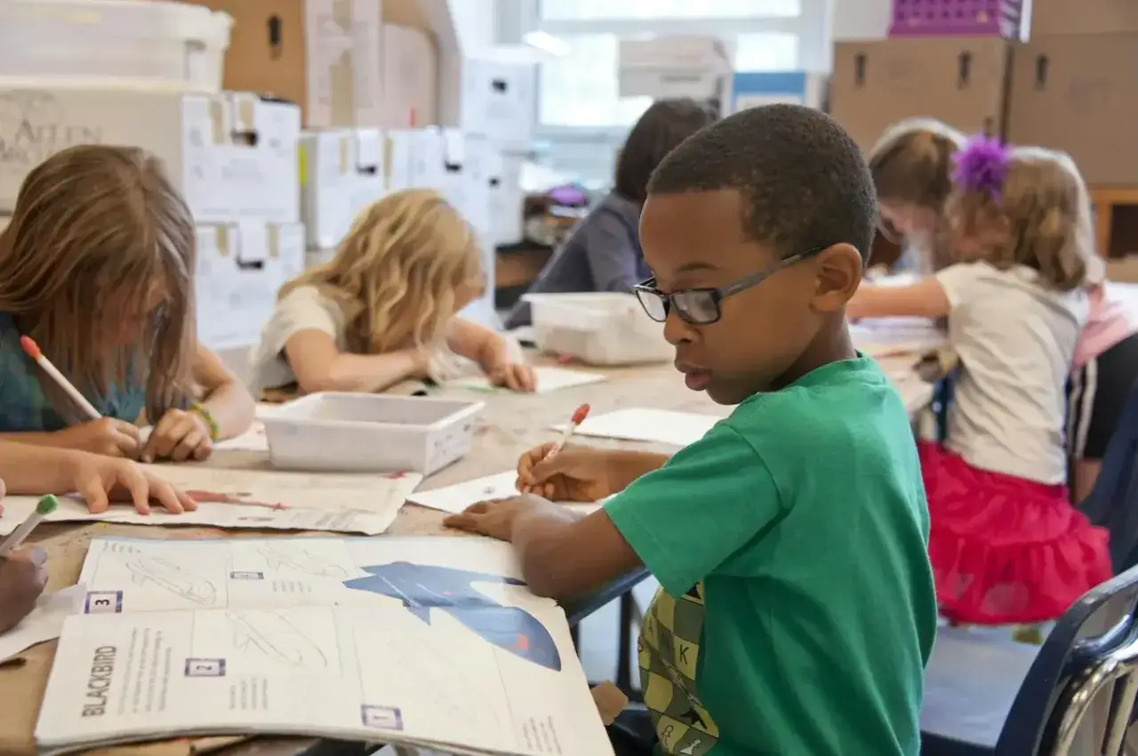 Kinder konzentriert bei einer Zeichenaufgabe. Ein Junge mit Brille und gr&uuml;nen T-Shirt ist im Vordergrund. Der Schulbegleiter hilft bei diesem Teil des Tagesablaufs.