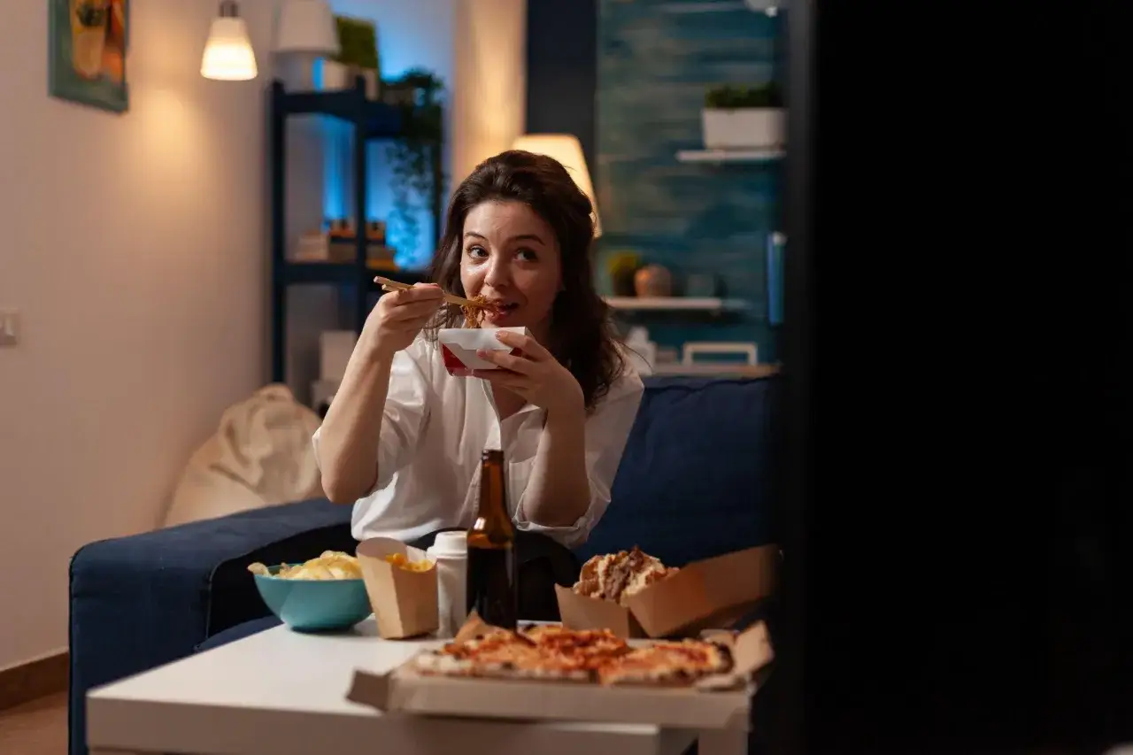 Mujer come fideos mientras ve TV, rodeada de comida rápida. Quizás por eso dice que dormir poco engorda.