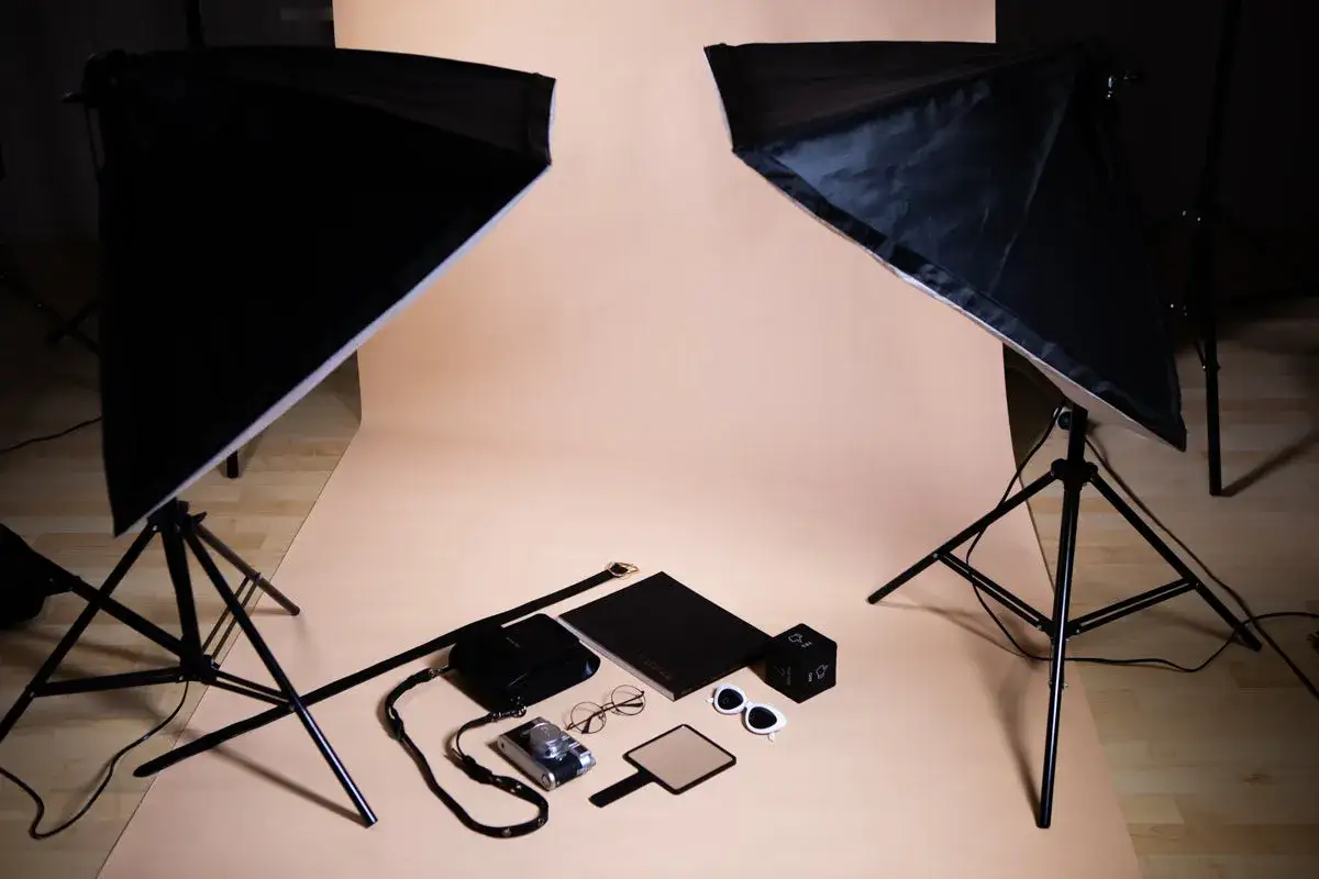 Flat lay z akcesoriami: aparat, torebka, okulary, lusterko, notes i kostka. Dwa softboxy oświetlają scenę.