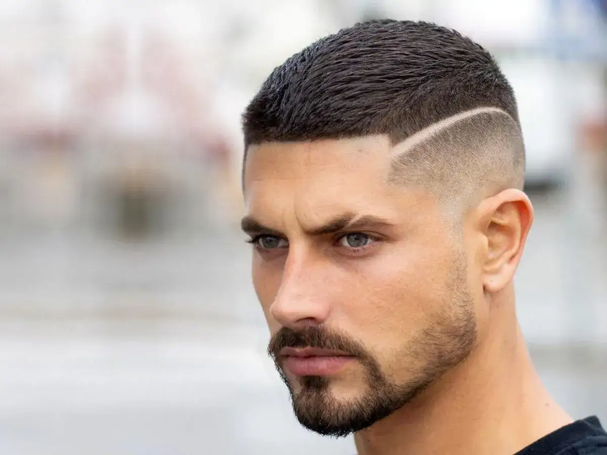 Uomo con capelli a spazzola corti, sfumati ai lati con una linea definita. Barba curata e sguardo intenso.