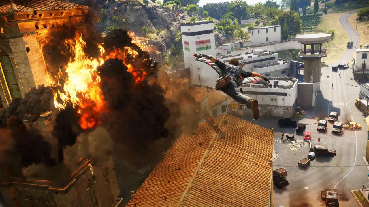 Rico z wingsuitem przelatuje nad wybuchem, uciekając przed wojskiem. Gra Just Cause 3 wymaga akcji!