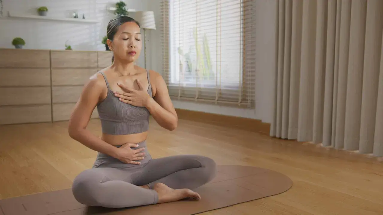 Kvinna sitter i lotusställning på en yogamatta, med slutna ögon och händerna på bröstet och magen, i en lugn stund av somatisk yoga.