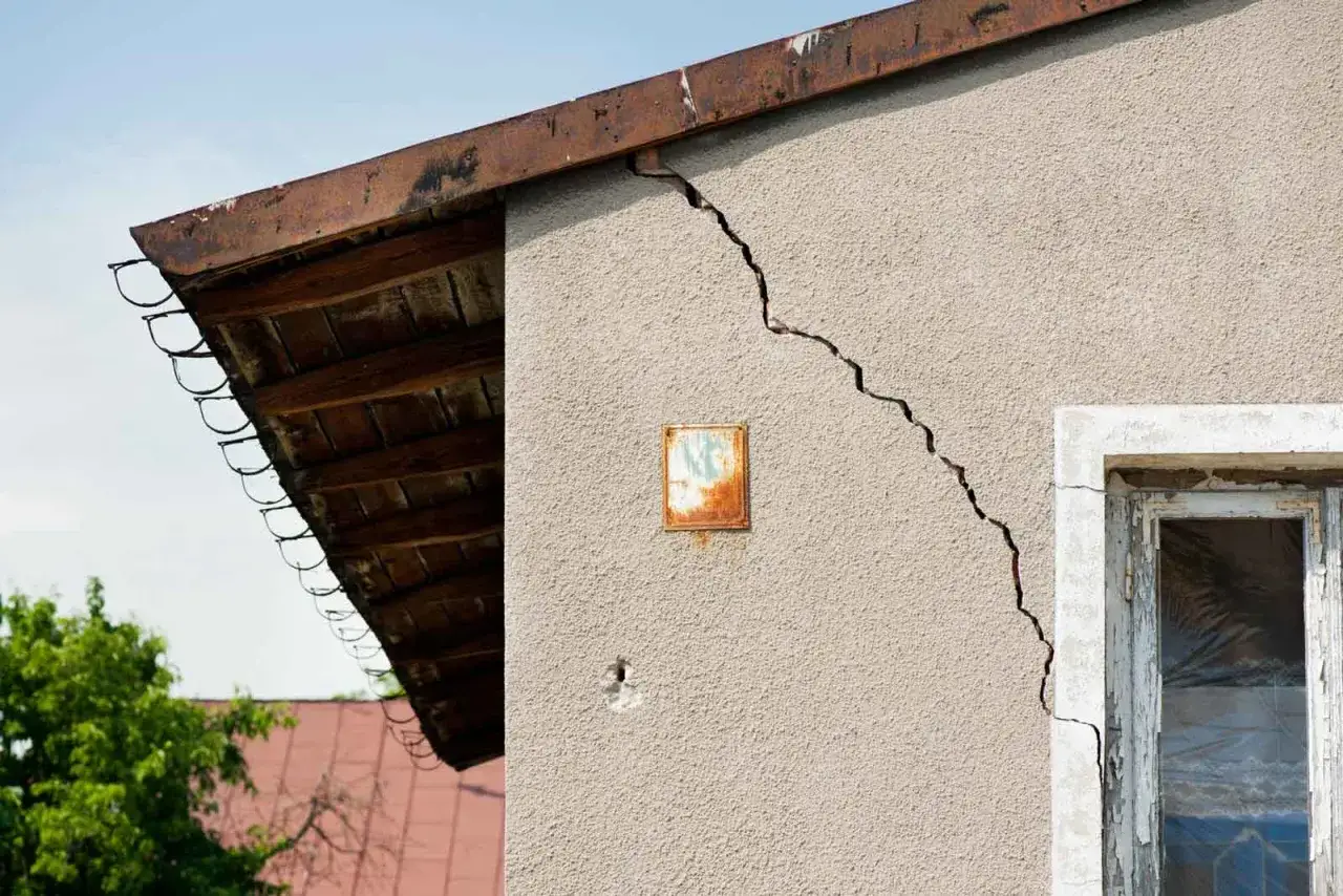 Grieta profunda en pared de edificio, comprometiendo la seguridad estructural. Se observa un alero de madera y un marco de ventana deteriorado.