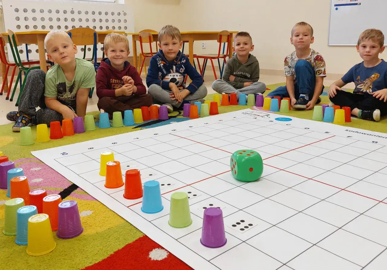 Dzieci siedzą wokół planszy do gry, przygotowując się do zabaw wyciszających w przedszkolu. Kolorowe kubeczki i zielona kostka czekają na ruch.